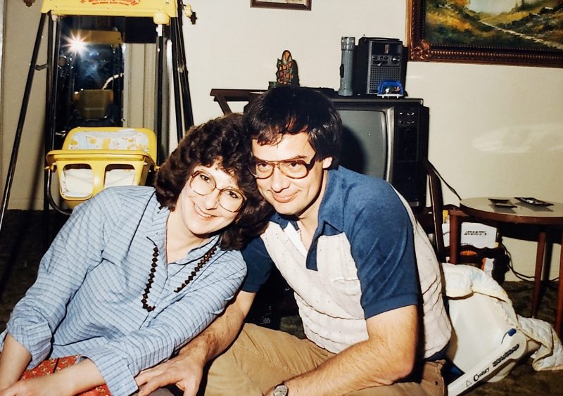 Laura and Daniel sit and smile together in their old home. Both are wearing 80's style glasses.