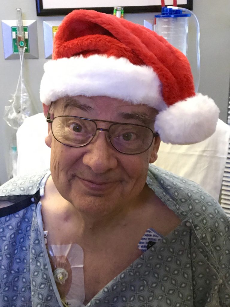 Daniel Hopkins wearing a santa hat and smiling in a hospital bed. Though the chemo port and hospital gown can be seen, his smile overpowers the situation he's in.