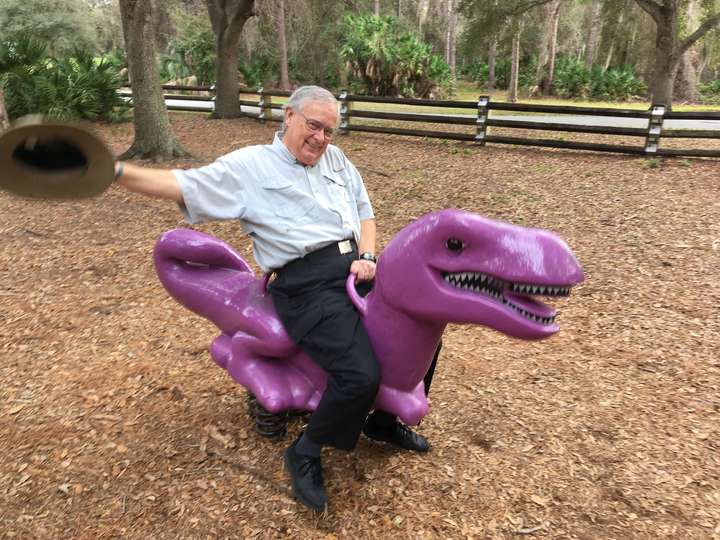 Dan Hopkins riding a purple dinosaur at a park and waving his hat in the hair.